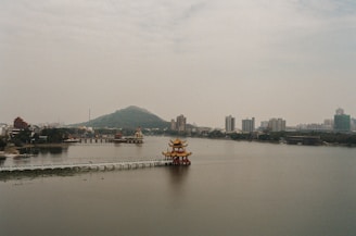 A serene lake with a traditional Chinese pavilion extending into the water. The backdrop features a cityscape with modern buildings and lush greenery. In the distance, a mountain looms under a cloudy sky, creating a peaceful and balanced scene.
