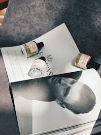 A flat lay composition featuring a monochrome magazine page with a close-up portrait of a man, partially covered by three small bottles of fragrance. The bottles, with labels visible, are arranged on a textured gray surface with sunlight casting soft shadows.