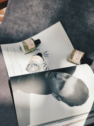 A flat lay composition featuring a monochrome magazine page with a close-up portrait of a man, partially covered by three small bottles of fragrance. The bottles, with labels visible, are arranged on a textured gray surface with sunlight casting soft shadows.