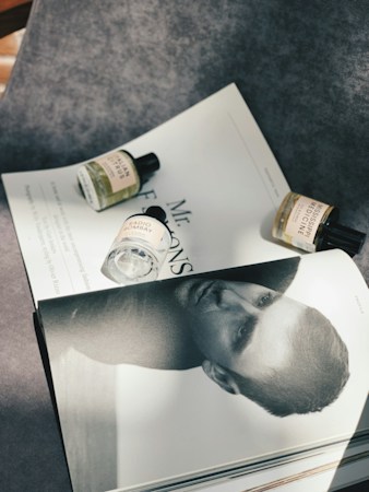 A flat lay composition featuring a monochrome magazine page with a close-up portrait of a man, partially covered by three small bottles of fragrance. The bottles, with labels visible, are arranged on a textured gray surface with sunlight casting soft shadows.