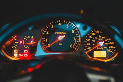 A close-up view of a car dashboard with illuminated gauges and warning lights. The speedometer, tachometer, and fuel gauge are prominently visible, displaying bright numbers and indicators against a dark background.