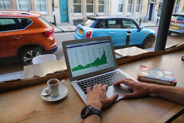 A person is working on a laptop at a wooden table next to a window. The screen displays a graph with rising data. Beside the laptop, there is a cup of espresso and a book titled 'How the World Thinks.' Outside the window, a street scene with parked cars can be seen.