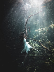 A woman in a flowing white dress is underwater, reaching towards a gnarled, sunlit branch. The scene is filled with an ethereal light filtering through the water, illuminating particles floating around. The underwater environment has a mystical quality, with greenish hues surrounding submerged branches and algae.
