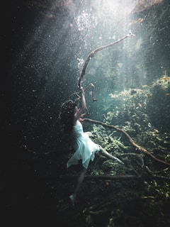 A woman in a flowing white dress is underwater, reaching towards a gnarled, sunlit branch. The scene is filled with an ethereal light filtering through the water, illuminating particles floating around. The underwater environment has a mystical quality, with greenish hues surrounding submerged branches and algae.