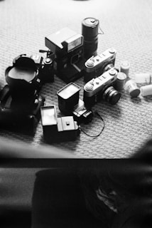 A collection of various cameras and photography equipment is scattered on a carpeted floor. The arrangement includes several vintage and modern cameras, film canisters, and camera lenses.