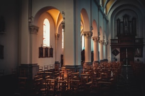 A quiet and dimly lit church interior featuring multiple rows of wooden chairs neatly arranged. Tall, arched windows allow soft, natural light to illuminate the stone columns and walls. On one side, religious paintings are displayed, adding to the reverent atmosphere. The dark wood of the organ pipes adds a classic touch at the far end of the aisle.