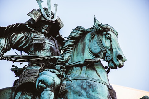 A bronze statue of a samurai warrior in full armor riding a horse. The samurai is wearing a traditional helmet with large, curved horns and detailed armor plates. The horse appears dynamic, captured mid-stride, with fine attention to its muscles and bridle.