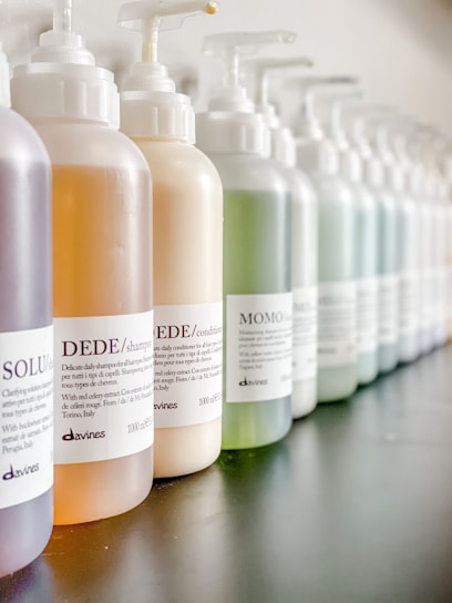 A close-up view of a row of various hair product bottles with pump dispensers arranged neatly. The bottles have white labels with black text, some featuring the brand 'Davines'. The bottles contain different colored liquids, indicating different products such as shampoo and conditioner.