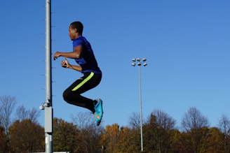 A person in athletic gear jumps high into the air against a clear blue sky. The scene is set in an outdoor area with visible trees and a lamp post in the background, suggesting a park or sports field environment.