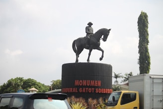 A statue of a mounted figure on a pedestal labeled 'Monumen Jenderal Gatot Soebroto' is prominently displayed in an outdoor setting. Several vehicles, including a blue car and a yellow truck, are seen around the statue. The background includes some greenery, trees, and a tall, cylindrical hedge or tree.