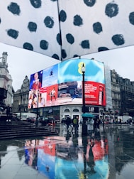 A city scene on a rainy day featuring large digital advertisements displayed on a building. The ground reflects the brightly lit screens due to the wet surface. People with umbrellas are walking around, and architectural structures surround the square.