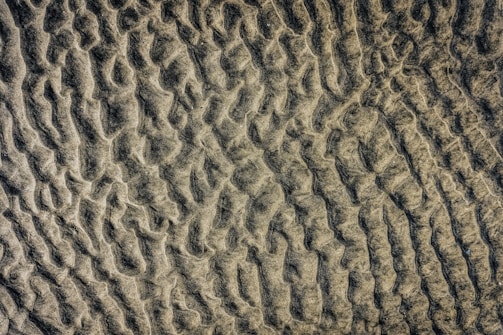Aerial view of a textured sandy surface with wave-like patterns and undulations. The sand appears to be slightly damp, creating a mixture of light and dark areas due to shadows and variations in the surface.