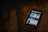 A smartphone displaying a search for a COVID-19 related app is placed on a dark wooden surface. The screen shows various app options with blue and white graphics.