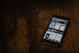 A smartphone displaying a search for a COVID-19 related app is placed on a dark wooden surface. The screen shows various app options with blue and white graphics.