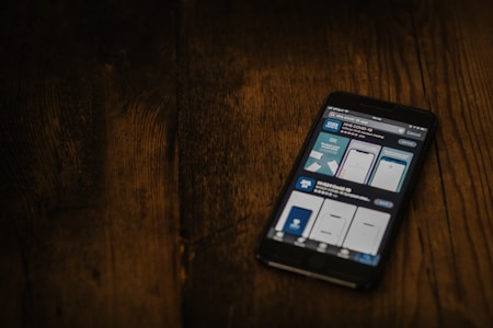A smartphone displaying a search for a COVID-19 related app is placed on a dark wooden surface. The screen shows various app options with blue and white graphics.