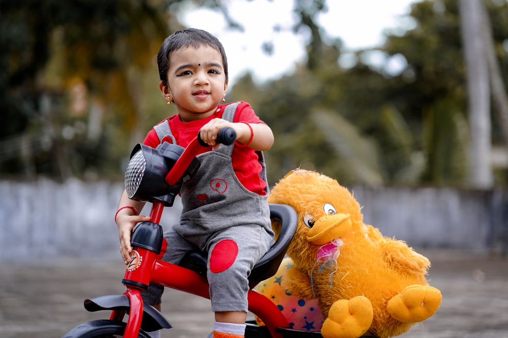 criança na jaqueta vermelha e branca que monta na bicicleta vermelha com o brinquedo de pelúcia do pato amarelo durante