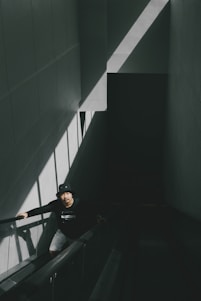 A person wearing a black hat and dark clothing ascends an escalator in a dimly lit environment. The scene is defined by stark contrasts between light and shadow with a triangular beam of sunlight illuminating the left wall.