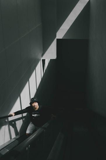 A person wearing a black hat and dark clothing ascends an escalator in a dimly lit environment. The scene is defined by stark contrasts between light and shadow with a triangular beam of sunlight illuminating the left wall.