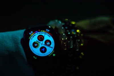 A smartwatch displays various health and activity metrics, including heart rate and activity rings, in a dimly lit environment. The screen is brightly lit with colorful graphics, providing a sharp contrast against the dark surroundings. The watch is worn on a wrist adorned with multiple beaded bracelets, adding a touch of personal style.