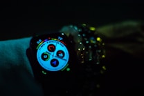 A smartwatch displays various health and activity metrics, including heart rate and activity rings, in a dimly lit environment. The screen is brightly lit with colorful graphics, providing a sharp contrast against the dark surroundings. The watch is worn on a wrist adorned with multiple beaded bracelets, adding a touch of personal style.
