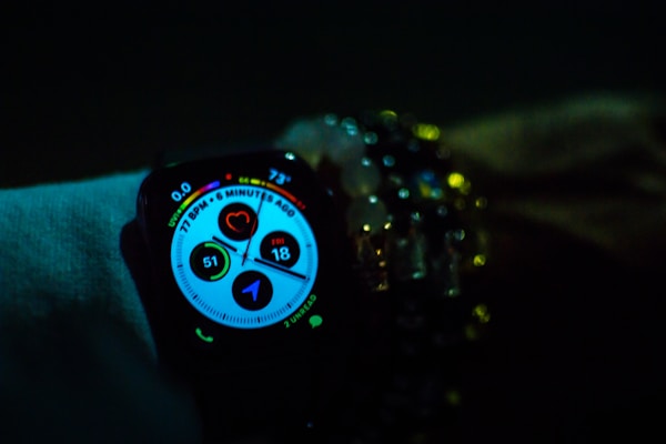 A smartwatch displays various health and activity metrics, including heart rate and activity rings, in a dimly lit environment. The screen is brightly lit with colorful graphics, providing a sharp contrast against the dark surroundings. The watch is worn on a wrist adorned with multiple beaded bracelets, adding a touch of personal style.