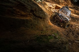 A remote-controlled off-road vehicle navigating a rugged, sandy terrain. The scene includes various textures of rocks and earth under dappled sunlight filtering through a forest or wooded area.