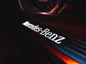 Close-up view of an illuminated Mercedes-Benz logo on what appears to be a car interior. The logo is glowing with white light against a dark background that includes shades of red and black.