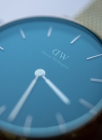 A close-up of a watch face with a minimalistic design, featuring a blue dial and white hour markers and hands. The brand name 'Daniel Wellington' is visible in the center of the dial.