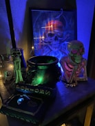 A collection of Halloween-themed items with a strong emphasis on skull imagery. There is a framed artwork of a skull wearing a bandana, illuminated with blue and orange lighting, creating an eerie atmosphere. On the table, a cauldron emits a mysterious green glow, surrounded by skeleton and skull figures. A potion book is visible, adding to the spooky ambiance.