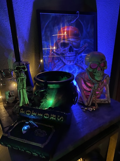 A collection of Halloween-themed items with a strong emphasis on skull imagery. There is a framed artwork of a skull wearing a bandana, illuminated with blue and orange lighting, creating an eerie atmosphere. On the table, a cauldron emits a mysterious green glow, surrounded by skeleton and skull figures. A potion book is visible, adding to the spooky ambiance.