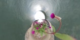 A floral arrangement is positioned at the center of a spiral or tunnel-like structure. Bright magenta flowers with green leaves are prominently visible, surrounded by a metallic-gray, swirling background that suggests a 360-degree perspective.