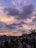 A picturesque residential neighborhood with palm trees and a series of rooftops stretches into the distance. Above, a dramatic sky filled with fluffy, dark clouds is painted in hues of orange, pink, and purple as the sun sets, casting a warm glow across the scene.