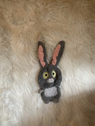 A plush toy resembling a rabbit with large yellow eyes, gray fur, and pink inner ears, is lying on a textured white surface resembling a fluffy rug.