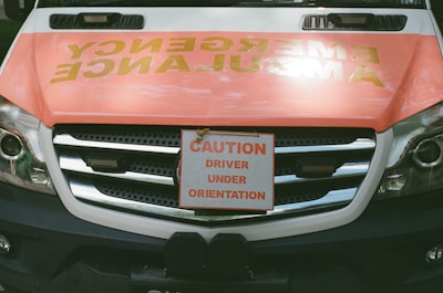 The front of an ambulance features a prominent sign attached to the grille that reads 'Caution Driver Under Orientation' in bold letters. The background shows the word 'Ambulance' written in reverse across the vehicle's hood in a high visibility color.
