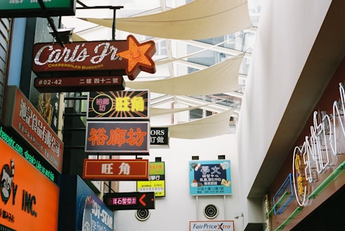 An indoor shopping area with multiple colorful, illuminated signs in various languages, including English and Chinese, hanging from the ceiling. The signs advertise food and other services. Overhead, the ceiling features a modern, partially open design with fabric sails for lighting and ventilation.