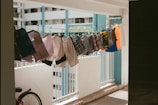 An array of various pieces of clothing is hung on a line within an apartment corridor, secured with colorful clothespins. The background shows a typical high-rise residential building with multiple windows, emphasizing an urban setting. A bicycle wheel is partially visible at the bottom left corner, adding to the everyday residential atmosphere.