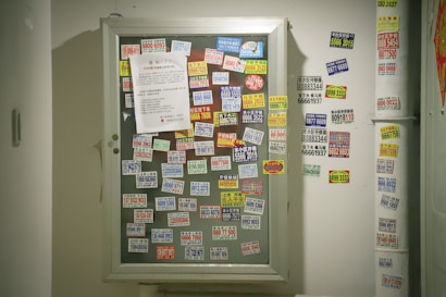 A bulletin board is covered with numerous brightly colored stickers displaying phone numbers and advertisements, primarily in Chinese text. A white paper is pinned amid the cluttered arrangement of stickers, which are randomly placed. Stickers are also visible on the adjacent wall, indicating heavy usage and layered application of advertisements.