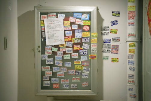 A bulletin board is covered with numerous brightly colored stickers displaying phone numbers and advertisements, primarily in Chinese text. A white paper is pinned amid the cluttered arrangement of stickers, which are randomly placed. Stickers are also visible on the adjacent wall, indicating heavy usage and layered application of advertisements.