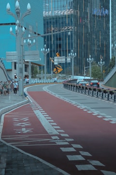 A modern city street featuring a red bike lane curving alongside a pedestrian path and road. Tall streetlights with bulbous top designs line the path, and skyscrapers made of glass and steel dominate the background. Cars are visible driving along, while a few pedestrians walk on the sidewalk. The overall setting suggests a busy urban environment.