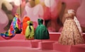 A group of vibrant and intricately designed dresses displayed on mannequins, set against a pink backdrop with abstract blurred lights. The dresses vary in color and texture, including a multi-colored floral gown, a bright orange coat, a lime green dress with black accents, a deep green gown, and a beige floral-patterned dress with lace details.