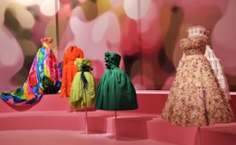 A group of vibrant and intricately designed dresses displayed on mannequins, set against a pink backdrop with abstract blurred lights. The dresses vary in color and texture, including a multi-colored floral gown, a bright orange coat, a lime green dress with black accents, a deep green gown, and a beige floral-patterned dress with lace details.