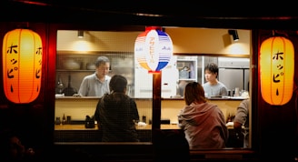 A cozy Japanese restaurant setting with two red lanterns on each side and a blue and red lantern in the middle. Inside, two chefs are working behind the counter, preparing food. Two customers are seated at the counter, engaging with the chefs.