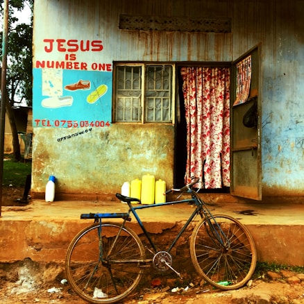 A small, weathered building with a hand-painted sign featuring shoes and the text 'Jesus is Number One' in red. A blue and yellow wall hosts a worn-out window and an open door with floral curtains inside. Several yellow jerry cans and a single white bottle sit in front of the building, along with a vintage bicycle leaning against the ledge.