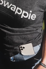 A person wearing a dark t-shirt with the word 'swappie' printed, partially tucked into blue denim jeans. A smartphone with multiple camera lenses is tucked into the pocket. The lower aspect of an arm and wrist with a woven bracelet is also visible.