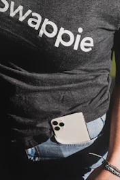 A person wearing a dark t-shirt with the word 'swappie' printed, partially tucked into blue denim jeans. A smartphone with multiple camera lenses is tucked into the pocket. The lower aspect of an arm and wrist with a woven bracelet is also visible.