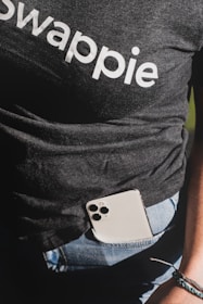 A person wearing a dark t-shirt with the word 'swappie' printed, partially tucked into blue denim jeans. A smartphone with multiple camera lenses is tucked into the pocket. The lower aspect of an arm and wrist with a woven bracelet is also visible.