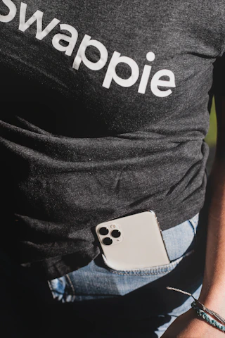 A person wearing a dark t-shirt with the word 'swappie' printed, partially tucked into blue denim jeans. A smartphone with multiple camera lenses is tucked into the pocket. The lower aspect of an arm and wrist with a woven bracelet is also visible.