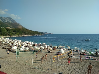 A sandy beach scene with numerous umbrellas and lounge chairs along the shore. People are sunbathing, playing beach volleyball, and enjoying the water. In the background, there is a calm sea with a few boats, and a mountainous landscape covered with greenery.