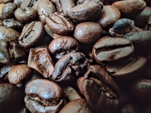 A close-up view of dark roasted coffee beans with a rich, oily surface texture. The beans are densely packed and vary slightly in size and shape, showcasing their deep brown color and pronounced creases.