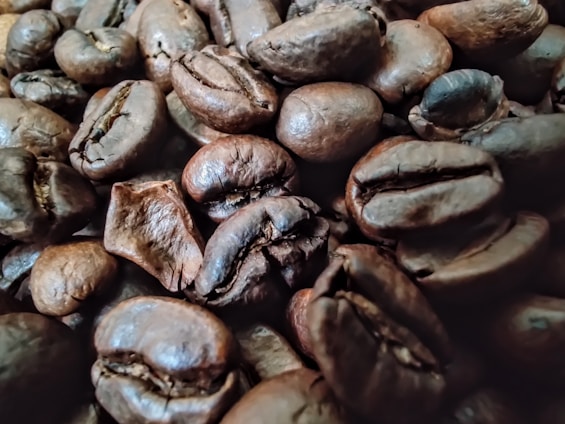 A close-up view of dark roasted coffee beans with a rich, oily surface texture. The beans are densely packed and vary slightly in size and shape, showcasing their deep brown color and pronounced creases.